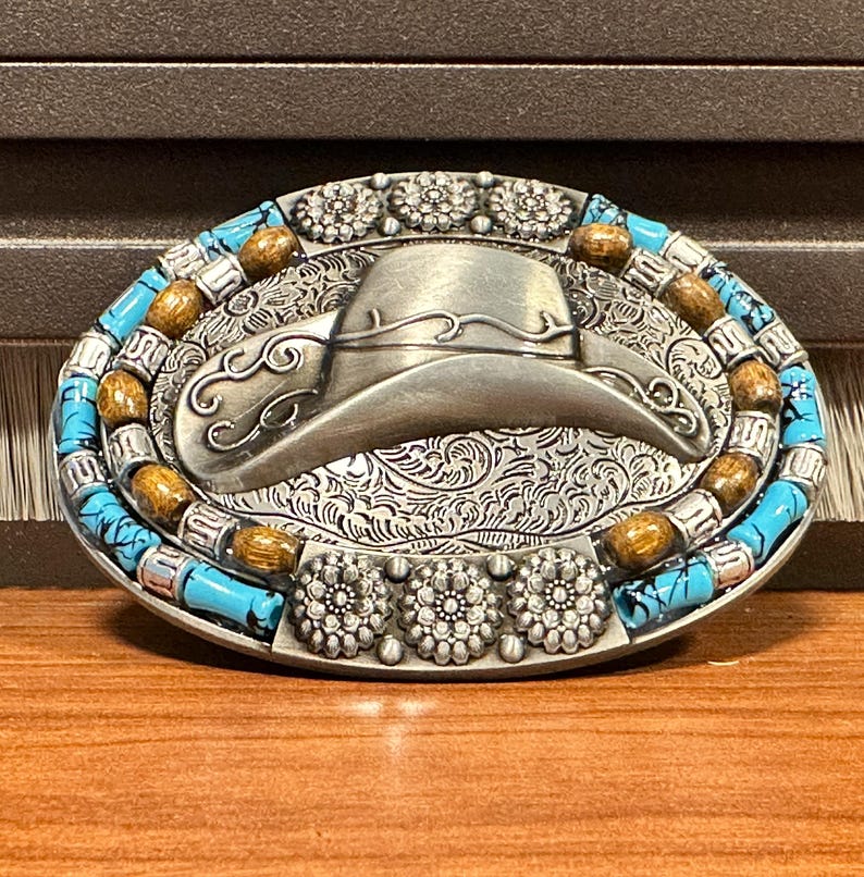 May include: Oval silver-tone belt buckle with a cowboy hat design. The buckle has turquoise and wooden bead accents around the border and floral details. The hat is in the centre of the buckle.