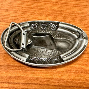 May include: Oval silver-coloured belt buckle with a cowboy boot design. The buckle has a detailed relief pattern and a sturdy metal loop for belt attachment. The buckle is made of metal.