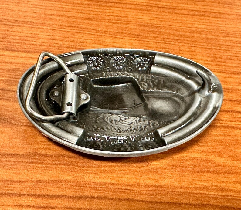 May include: Oval silver-coloured belt buckle with a cowboy boot design. The buckle has a detailed relief pattern and a sturdy metal loop for belt attachment. The buckle is made of metal.