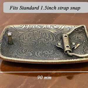 May include: A gold-toned belt buckle with an ornate floral design. It measures 3.5 inches by 1.5 inches and is labeled "Fits Standard 1.5 inch strap snap".