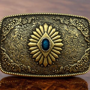 May include: A gold-toned belt buckle with an ornate design featuring a blue gemstone in the center. The buckle has a detailed floral pattern around the edges.