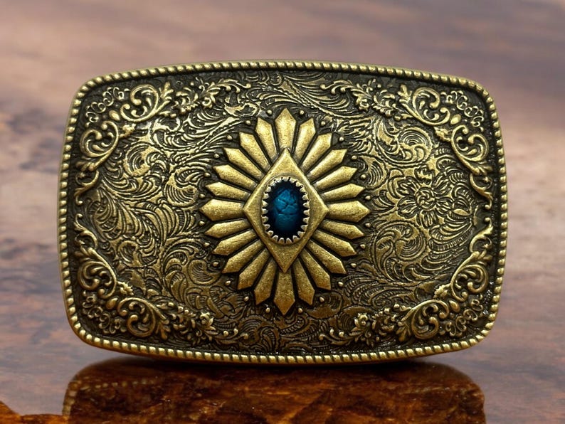 May include: A gold-toned belt buckle with an ornate design featuring a blue gemstone in the center. The buckle has a detailed floral pattern around the edges.