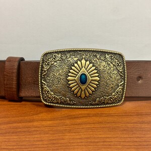 May include: A brown leather belt with a gold-toned buckle featuring an ornate design and a blue gemstone in the center.