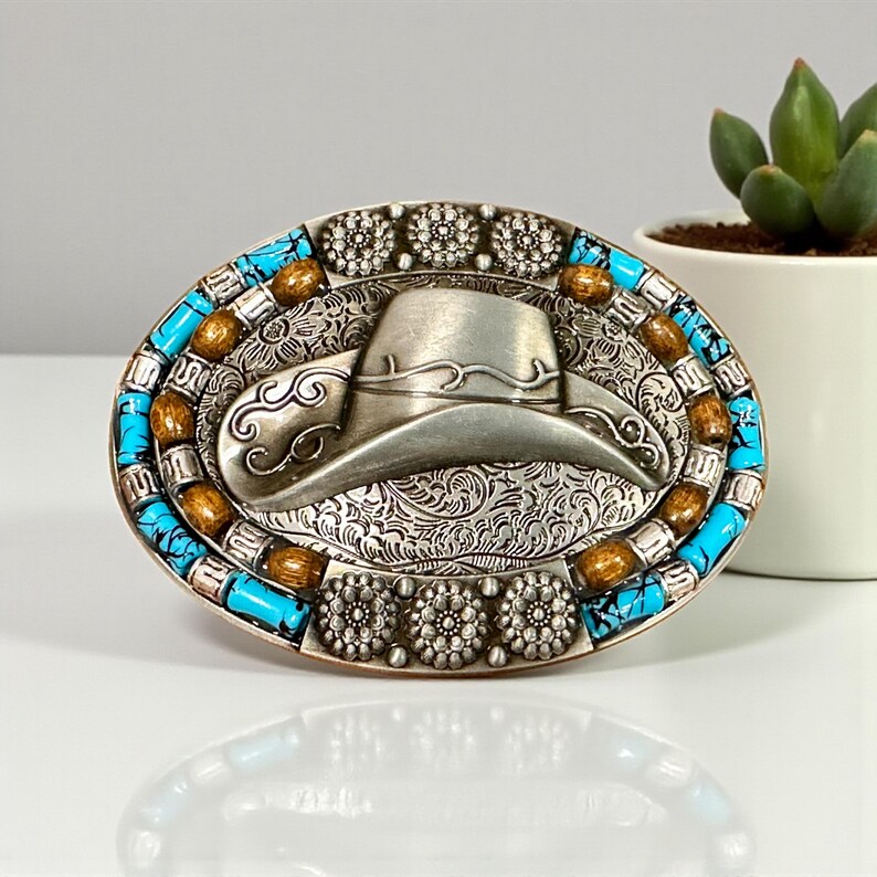 May include: An oval silver-tone belt buckle with a cowboy hat design. The buckle is decorated with turquoise and brown stones around the edge and floral patterns. A small succulent plant in a white pot is in the background.