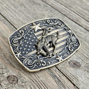 May include: A rectangular, antique brass-colored belt buckle featuring a rodeo scene. The buckle has a design of a cowboy riding a bucking bronco against a backdrop of the American flag. Decorative scrollwork and stars frame the design.