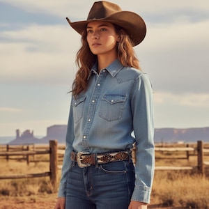May include: A woman wearing a brown cowboy hat, denim shirt, and blue jeans. The shirt has two chest pockets and pearl snap buttons. A decorative belt with a silver buckle and patterned leather is also visible. The background features a wooden fence and a desert landscape.