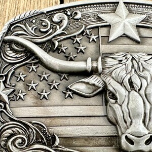 May include: Silver-toned metal belt buckle featuring a longhorn steer head and a star-studded American flag design. The buckle has a detailed, embossed pattern with a rope-like border and decorative flourishes.