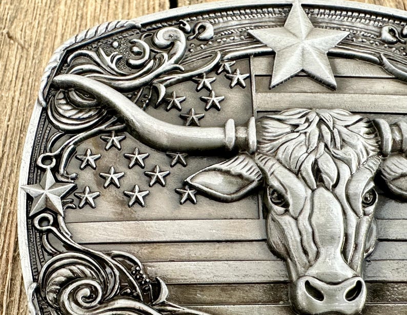 May include: Silver-toned metal belt buckle featuring a longhorn steer head and a star-studded American flag design. The buckle has a detailed, embossed pattern with a rope-like border and decorative flourishes.
