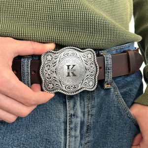 May include: A silver belt buckle with an ornate design and a circular center featuring the letter "K" and the name "Kevin" engraved on it. The buckle is attached to a brown leather belt.