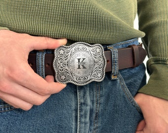 Puede incluir: Una hebilla de cinturón de plata con un diseño ornamentado y un centro circular que presenta la letra "K" y el nombre "Kevin" grabado en él. La hebilla está unida a un cinturón de cuero marrón.