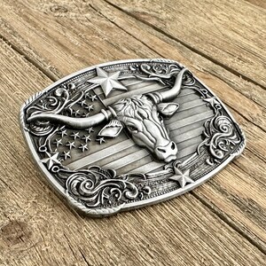 May include: Silver-toned metal belt buckle featuring a longhorn steer head with large horns, set against a backdrop of the American flag. The buckle has a rectangular shape with decorative scrollwork and stars around the edges, evoking a Western theme.