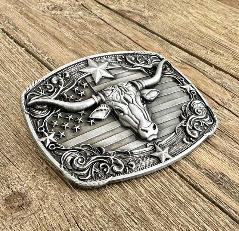 May include: Silver-toned metal belt buckle featuring a longhorn steer head with large horns, set against a backdrop of the American flag. The buckle has a rectangular shape with decorative scrollwork and stars around the edges, evoking a Western theme.