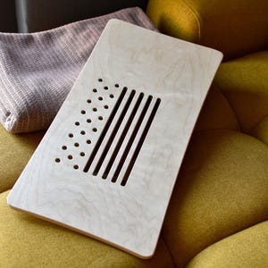 Può includere: Un supporto per laptop in legno chiaro con un design a taglio che presenta una serie di fessure orizzontali e una griglia di piccoli fori. Il supporto è appoggiato su una superficie di tessuto giallo.