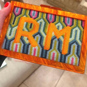 Può includere: Una pochette rettangolare con una cornice in acrilico marmorizzato arancione brillante. Il pannello centrale presenta un ricamo con un motivo geometrico nei toni del blu, verde e rosa, sormontato dalle grandi lettere arancioni ricamate RM.