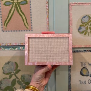 Puede incluir: Un clutch rectangular rosa con apariencia veteada. El clutch tiene un interior de malla blanca y un pequeño cierre rosa a juego. Se ven brazaletes dorados en la muñeca que sostiene el clutch. Arte de punto de aguja en el fondo.