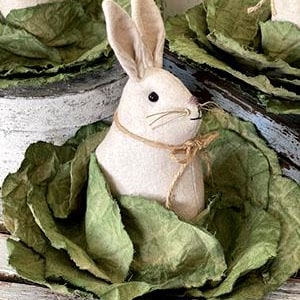 May include: A white felt bunny rabbit with brown eyes and a brown felt nose is sitting in a green cabbage. The cabbage is made of fabric and has a rustic, vintage look.