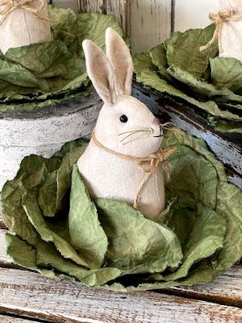 May include: A white felt bunny rabbit with brown eyes and a brown felt nose is sitting in a green cabbage. The cabbage is made of fabric and has a rustic, vintage look.