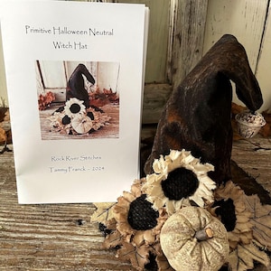May include: A black witch hat with brown and white sunflower and pumpkin embellishments. The hat is sitting on a wooden surface. The text on the card reads "Primitive Halloween Neutral Witch Hat" and "Rock River Stitches Tammy Franck - 2024".