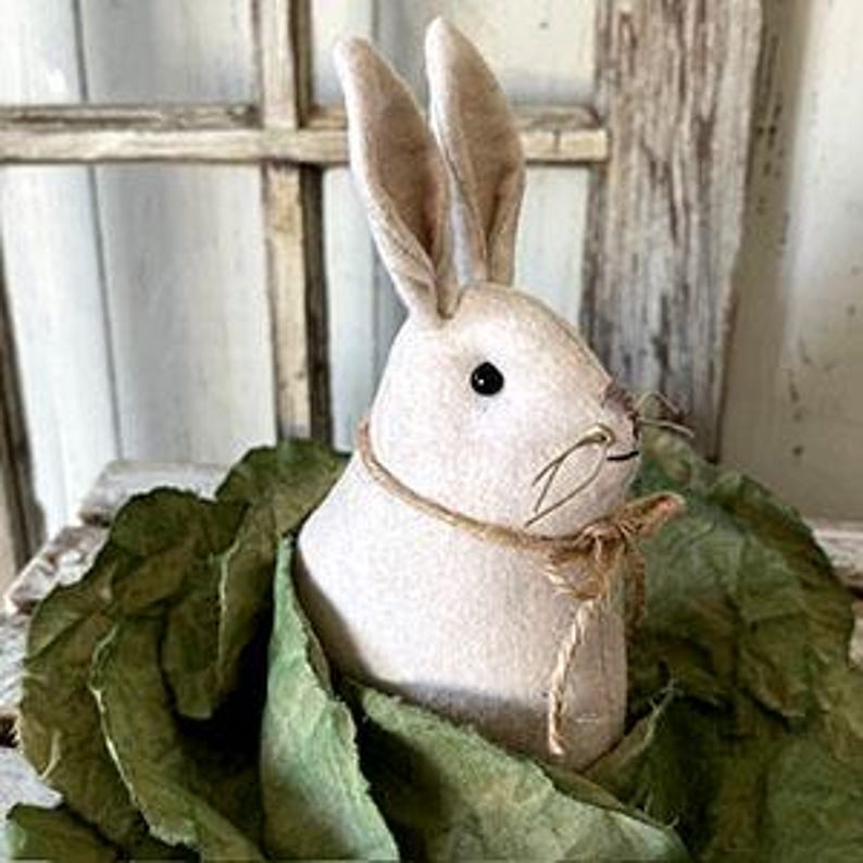 May include: A white felt bunny with brown eyes and a hessian bow around its neck. The bunny is sitting in a bed of green felt leaves.