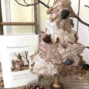 Puede incluir: Un patrón de artesanía de árbol de hojas de otoño primitivo con una base de arpillera marrón y un libro titulado "Primitive Fall Leaves Tree" de Rock River Stitches, Tammy Franck - 2021.