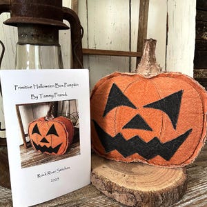 Pode incluir: Uma abóbora de Halloween de tecido laranja com um rosto de jack-o'-lantern esculpido repousa sobre uma fatia de madeira. Um cartão próximo diz "Primitive Halloween Box Pumpkin By Tammy Franck" e "Rock River Stitches 2023". Uma lanterna vintage está ao fundo.