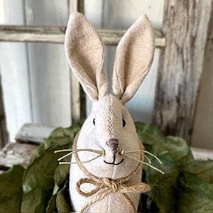 May include: A beige felt bunny rabbit with brown eyes and a brown nose. The bunny has a brown twine bow tied around its neck and brown whiskers.