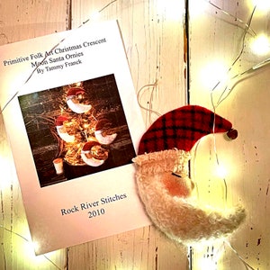 May include: A book titled "Primitive Folk Art Christmas Crescent Moon Santa Ornies by Tammy Franck" with a photo of a Christmas tree decorated with crescent moon ornaments. The book is published by Rock River Stitches in 2010.