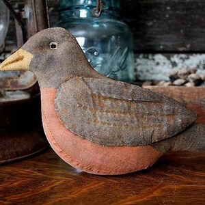 Puede incluir: Un pájaro de tela hecho a mano en forma de petirrojo con un cuerpo gris y marrón y un pecho rojo. El pájaro está sentado sobre una superficie de madera.