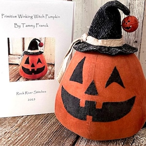 Peut inclure: Une décoration en forme de citrouille orange avec un visage souriant noir et un chapeau de sorcière noir. Le chapeau a un bandeau beige et un petit ornement de citrouille. Le texte sur la carte indique "Primitive Winking Witch Pumpkin By Tammy Franck" et "Rock River Stitches 2023".