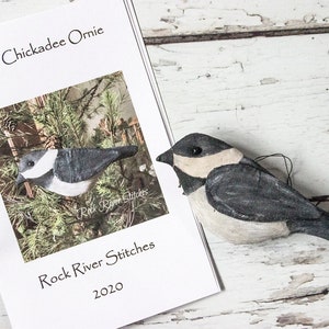 Könnte beinhalten: Ein weißes Papiermusterheft mit dem Titel "Primitive Chickadee Ornie" und dem Text "Rock River Stitches 2020". Neben dem Heft liegt ein kleines, handgenähtes, grau-weißes Meisen-Ornament.