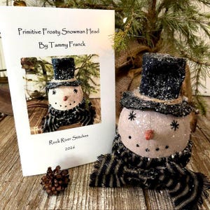 May include: A snowman head figurine wearing a black top hat and a black and tan striped scarf, accompanied by a printed instruction card titled Primitive Frosty Snowman Head By Tammy Franck, Rock River Stitches 2026, and a pinecone.