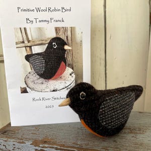 May include: A black and gray wool robin bird with an orange breast. The bird is sitting on a white surface. The image includes the text "Primitive Wool Robin Bird By Tammy Franck Rock River Stitches 2023".