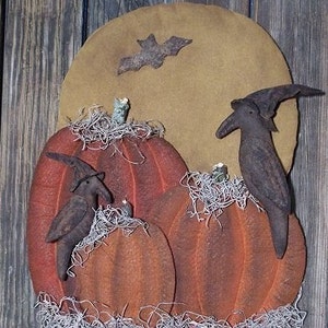Puede incluir: Tres calabazas naranjas con tallos marrones y una textura marrón y peluda están dispuestas frente a una luna redonda marrón con una silueta de murciélago negro. Dos cuervos negros con sombreros de bruja están posados ​​en las calabazas.