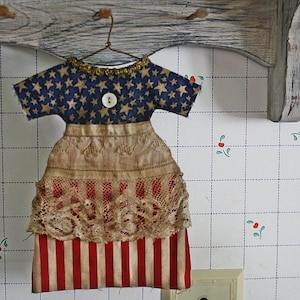 Op de afbeelding: Een stoffen jurk ornament in patriottische kleuren. De bovenkant is marineblauw met gouden sterren, het midden is beige kant en de onderkant is rood en wit gestreept. Het hangt aan een draadkleerhanger.