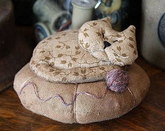 May include: A brown and tan fabric cat with a ball of yarn, sleeping on a brown fabric bed.