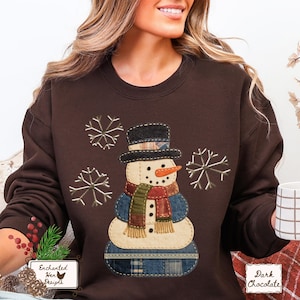 May include: Dark chocolate brown sweatshirt featuring a snowman applique with a black top hat, orange carrot nose, and a colorful scarf. The snowman is surrounded by embroidered snowflakes. The text "Enchanted Hen Designs" is on a small tag.