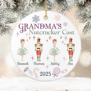 Może przedstawiać: Biała ceramiczna ozdoba świąteczna z napisem "GRANDMA'S Nutcracker Cast" w kolorze fioletowym i zielonym. Ozdoba zawiera ilustracje baletnic i żołnierzyków do orzechów, z imionami i rokiem 2025.