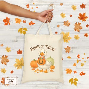 May include: Cream-colored tote bag with the text "Honk or Treat" and a cartoon duck holding a jack-o'-lantern bucket. The bag features pumpkins and candy corn, perfect for Halloween. The bag is held by a person's hand.
