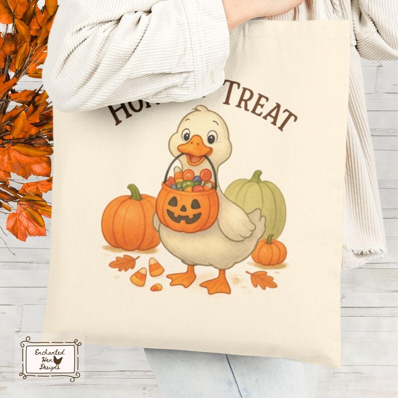 May include: Cream-colored tote bag featuring a cartoon duck holding a jack-o'-lantern candy bucket. The bag has the words "Hunt Treat" in brown. Pumpkins and fall leaves surround the duck, creating a Halloween theme.