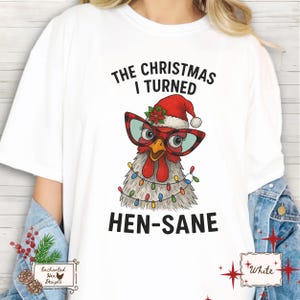 May include: White t-shirt with a Christmas-themed graphic. The graphic features a cartoon chicken wearing a Santa hat, glasses, and Christmas lights, with the text "THE CHRISTMAS I TURNED HEN-SANE."