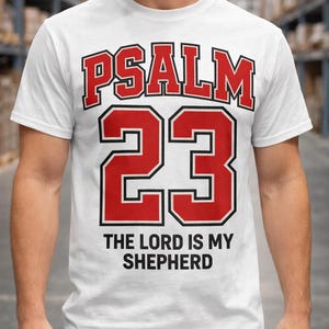 Puede incluir: Una camiseta blanca de manga corta con el texto rojo PSALM 23 en una fuente atlética negrita sobre un número 23 grande de color rojo con borde negro. Debajo del número, el texto negro dice THE LORD IS MY SHEPHERD en una fuente sans-serif.