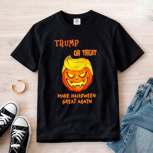 Puede incluir: Una camiseta negra de manga corta con un gráfico de una calabaza de Halloween con pelo amarillo. El texto TRUMP OR TREAT aparece sobre la calabaza y MAKE HALLOWEEN GREAT AGAIN debajo en letras naranjas. Tiene cuello redondo.