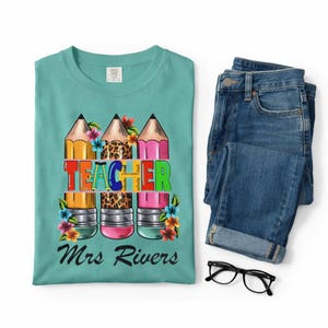 Puede incluir: Una camiseta de color verde azulado con un gráfico de tres lápices con flores coloridas y la palabra TEACHER en letras multicolores, con el nombre Mrs Rivers impreso debajo. Junto a la camiseta hay unos vaqueros azules y gafas negras.