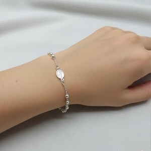 Könnte beinhalten: Ein silbernes Rosenkranzarmband mit kleinen, runden Perlen und einem ovalen Anhänger mit einer religiösen Figur. Das Armband wird an einem Handgelenk getragen.