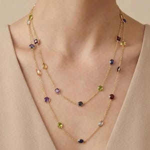 Può includere: Una collana a doppia catena in oro con una varietà di pietre preziose multicolori in diverse forme, tra cui tagli rotondi, ovali e quadrati. Le pietre sono posizionate a intervalli lungo le delicate maglie dorate della catena.