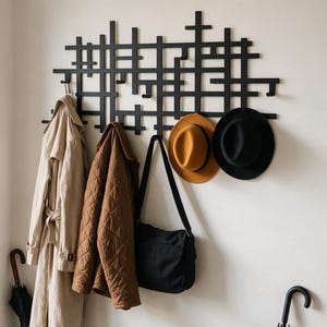 May include: A black metal wall-mounted coat rack with a grid-like geometric design featuring multiple hooks. Hanging from the rack are a beige trench coat, a brown quilted jacket, a black messenger bag, a mustard yellow hat, and a black hat.