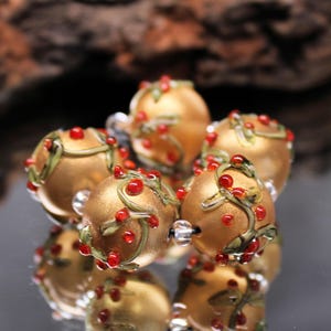May include: Close-up of several gold-colored glass beads. Each bead is adorned with red berries and green vines. The beads are arranged in a circular pattern, with a reflective surface below.