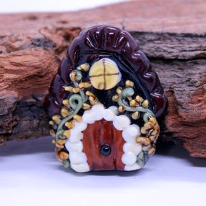 May include: A handcrafted glass bead shaped like a small house with a dark maroon roof, a brown door framed by white dots, a yellow circular window, and green vine-like details with small yellow accents on a black background.