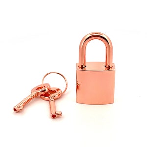 May include: A rose gold padlock with two matching keys. The padlock has a square body and a rounded top.