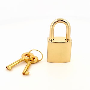May include: A gold padlock with two matching keys. The padlock has a square body and a rounded top.